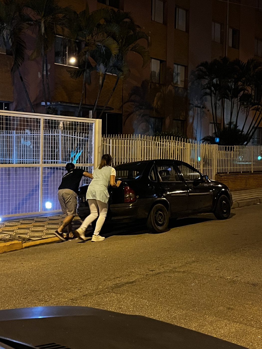 Duas pessoas empurrando um carro preto em uma rua, à noite